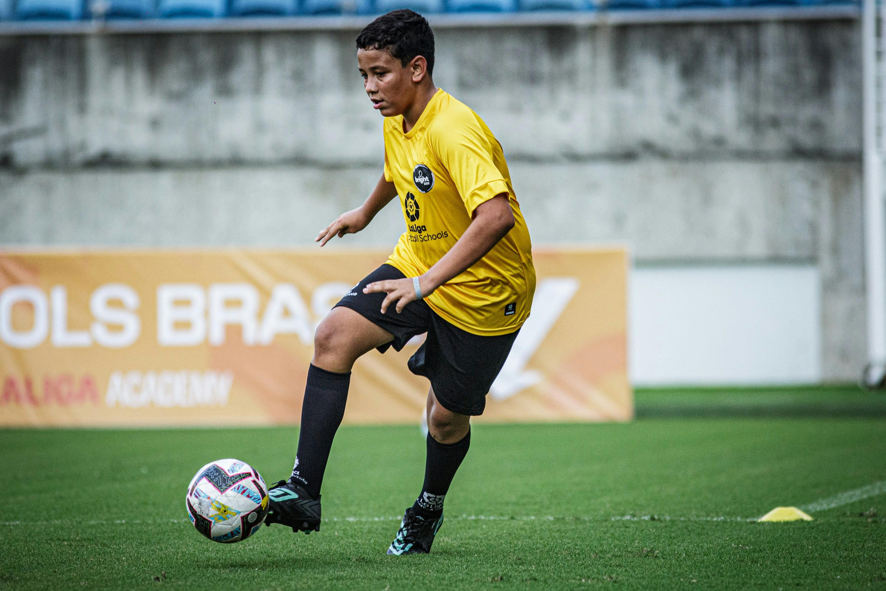 Garoto vestido com uniforme da LALIGA Academy Schools Brasil em estadio de futebol, jogando futebol